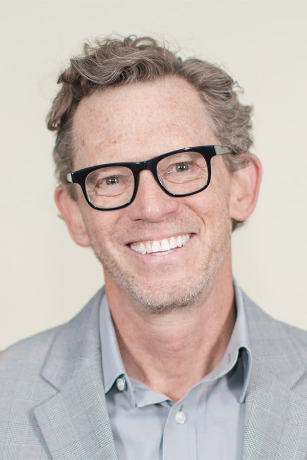 A smiling man with curly graying hair, dark-framed glasses, and a light gray blazer over a blue collared shirt.