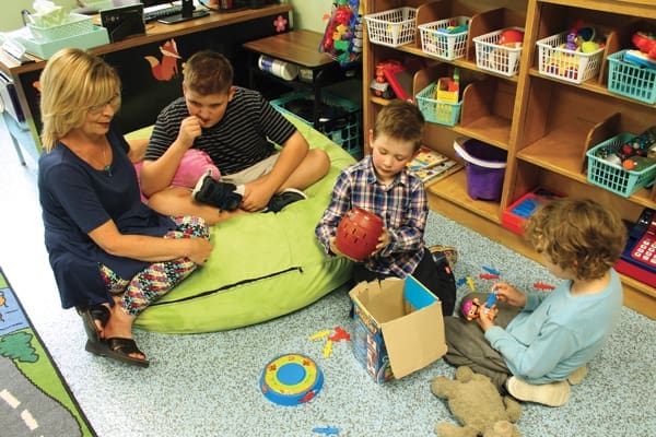 A woman sits on a green bean bag with two boys playing with toys on a classroom floor.