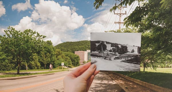 A hand holds a black-and-white historic photo over a sunny street with a cyclist and a brick building.