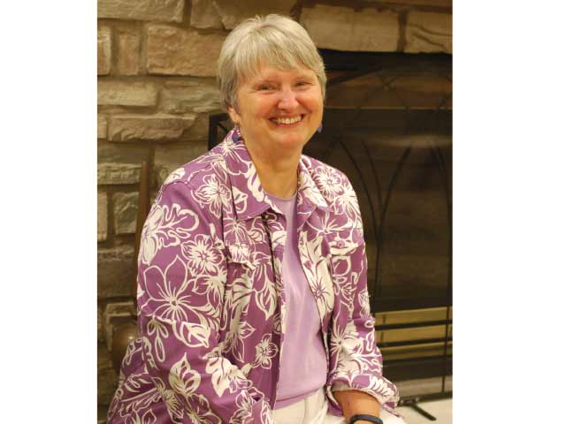 A smiling woman with short gray hair in a purple floral jacket, seated before a stone fireplace.