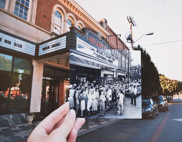 A hand holds a vintage black-and-white photo of a crowd outside the Grandin Theatre over the same street today.