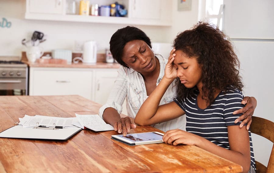 A woman with her hand on a stressed teenage girl's shoulder as they look at a tablet at a wooden kitchen table.