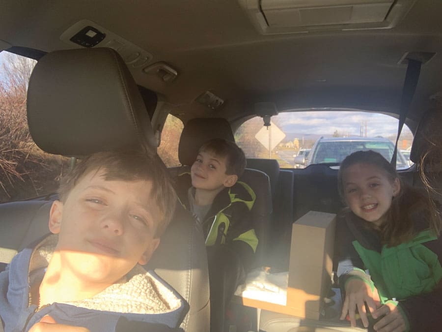 Three children sit in the back seat of a car, smiling and squinting in bright sunlight, with a cardboard box beside them.