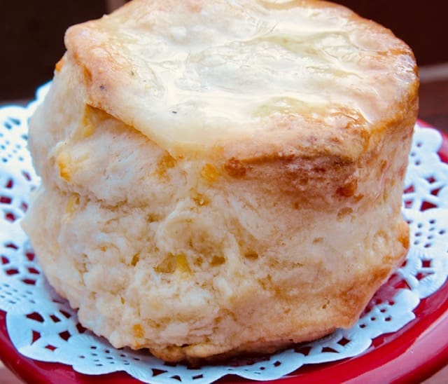 A tall, golden cheese biscuit with a glossy buttered top, sitting on a white doily atop a red plate.