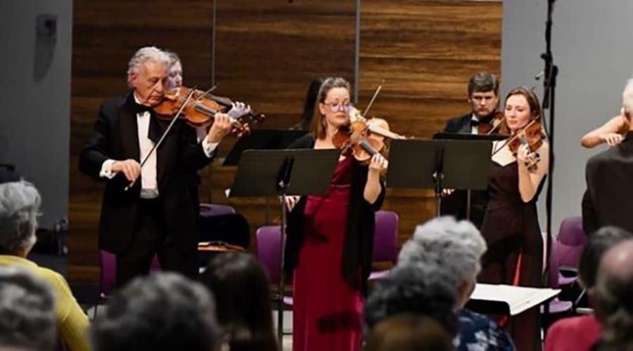 A white-haired violinist in a tuxedo performs at the front of a string ensemble before a seated audience.