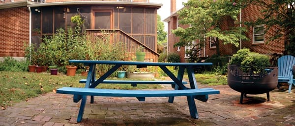 A blue wooden picnic table on a brick patio beside a screened porch, with a black fire pit and potted plants.