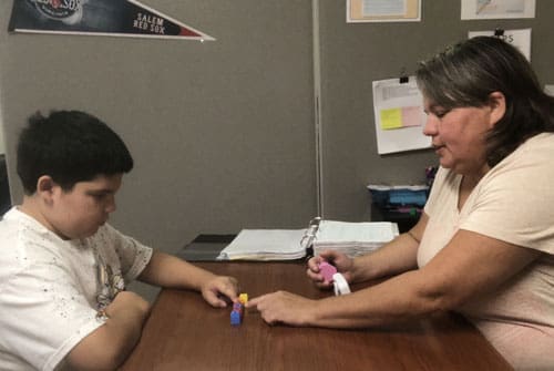 A young man stacks colorful blocks on a wooden table while a woman in a pale top guides him.