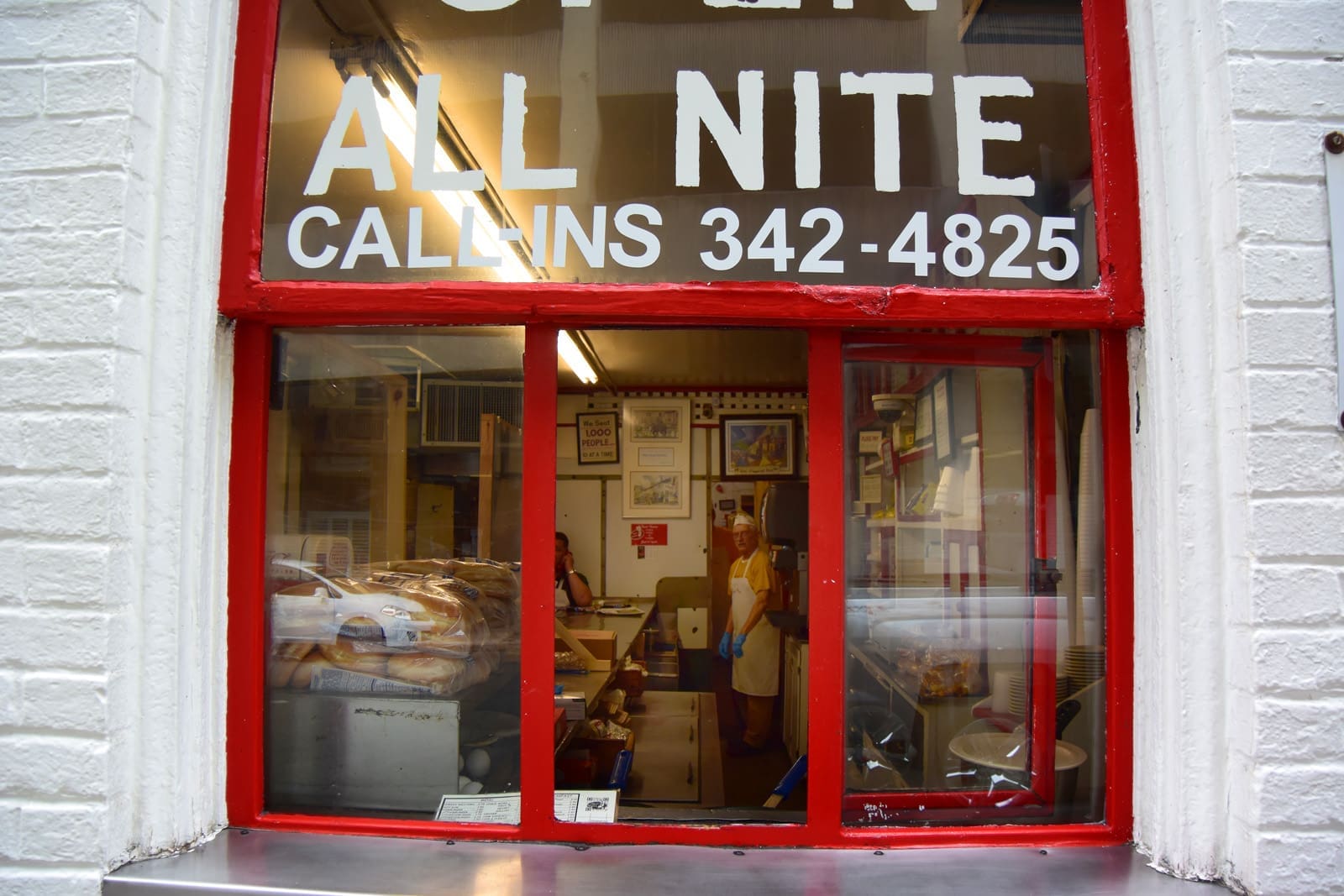 Red-framed bakery window with a sign reading "ALL NITE" and "CALL-INS 342-4825," with a baker in an apron inside.