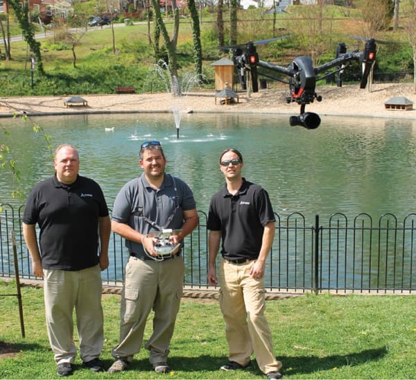 Three men in black polo shirts stand by a pond as a black drone with a camera hovers overhead, one holding a controller.