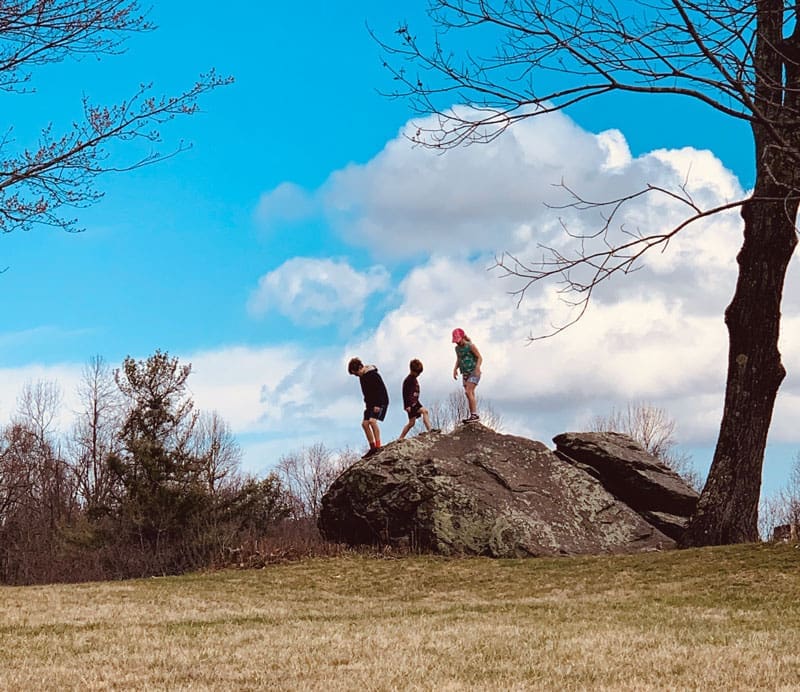 Three kids climb and stand on a large lichen-covered boulder under a bright blue, cloudy sky, with bare trees.