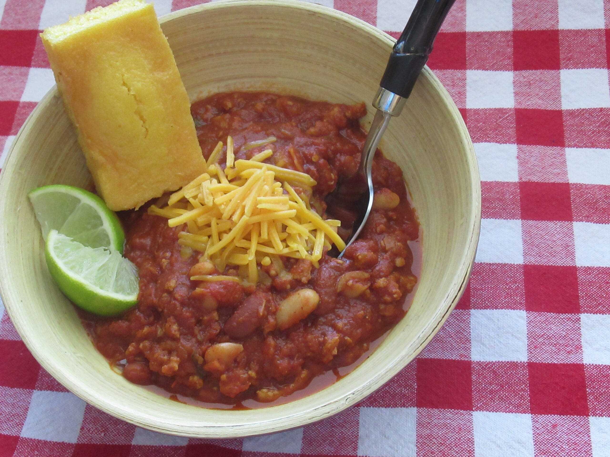 Bowl of red chili with beans, shredded cheddar, a lime wedge and cornbread on a red gingham tablecloth.