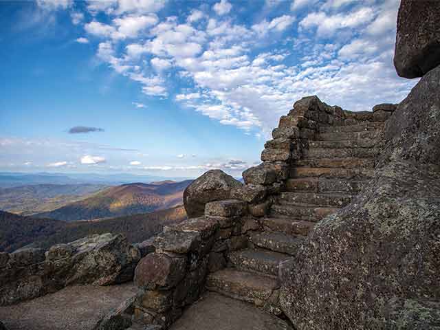Worn stone steps climb a rocky outcrop above layered blue mountain ridges under a partly cloudy sky.