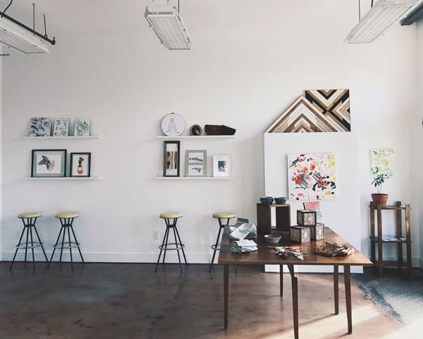 A bright gallery room with white walls, framed prints on shelves, yellow-topped stools, and a wooden table with ceramics.
