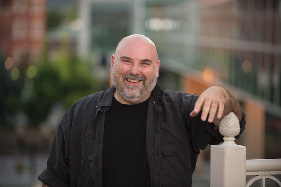 A smiling bald man with a gray beard leans on a white railing post outdoors, wearing a black shirt.