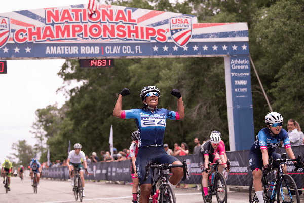 A cyclist in a blue jersey with race number 24 raises both fists as she crosses the finish line at the National Championships.