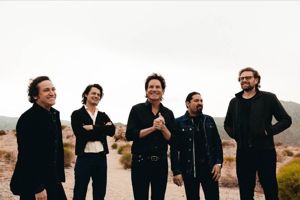 Five men in dark clothing stand smiling on a desert hillside under a pale sky.