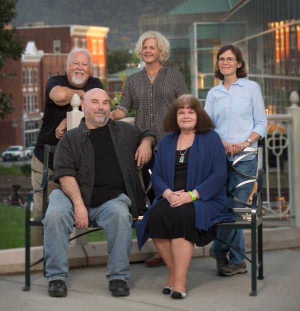 Five people posing outdoors on a patio near brick buildings at dusk, two seated on a bench and three standing behind.