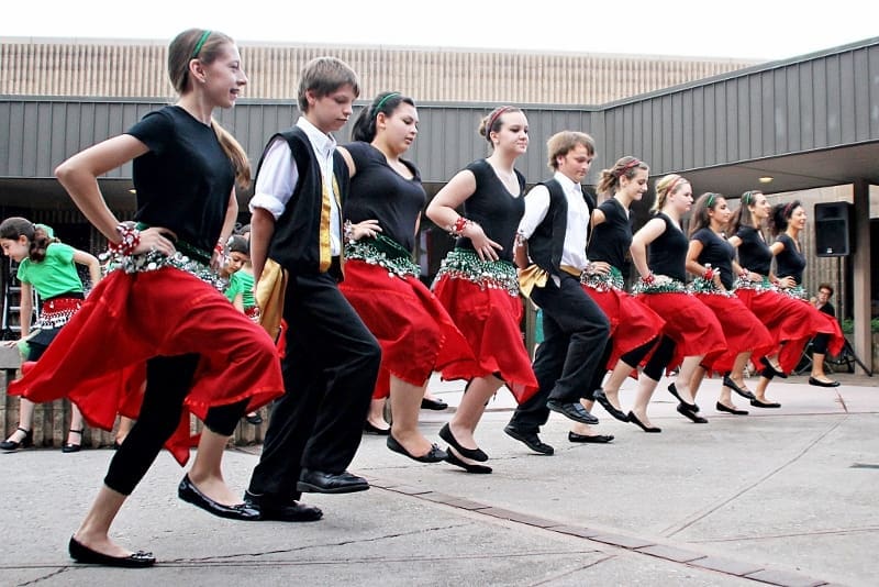 A line of young dancers in black tops, red skirts and black vests performs a synchronized folk dance outdoors.