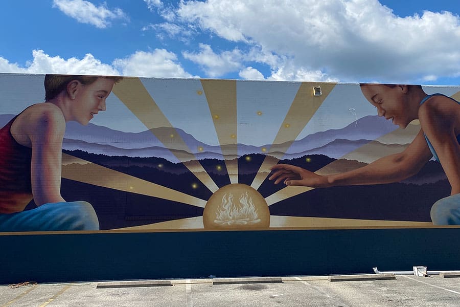 A large outdoor mural shows two young people reaching toward a glowing sun over blue mountain ridges, with golden rays and a campfire.