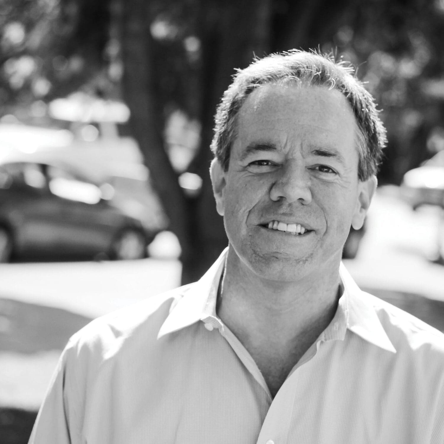 A smiling man in an open-collared shirt stands outdoors before blurred trees and parked cars, in black and white.