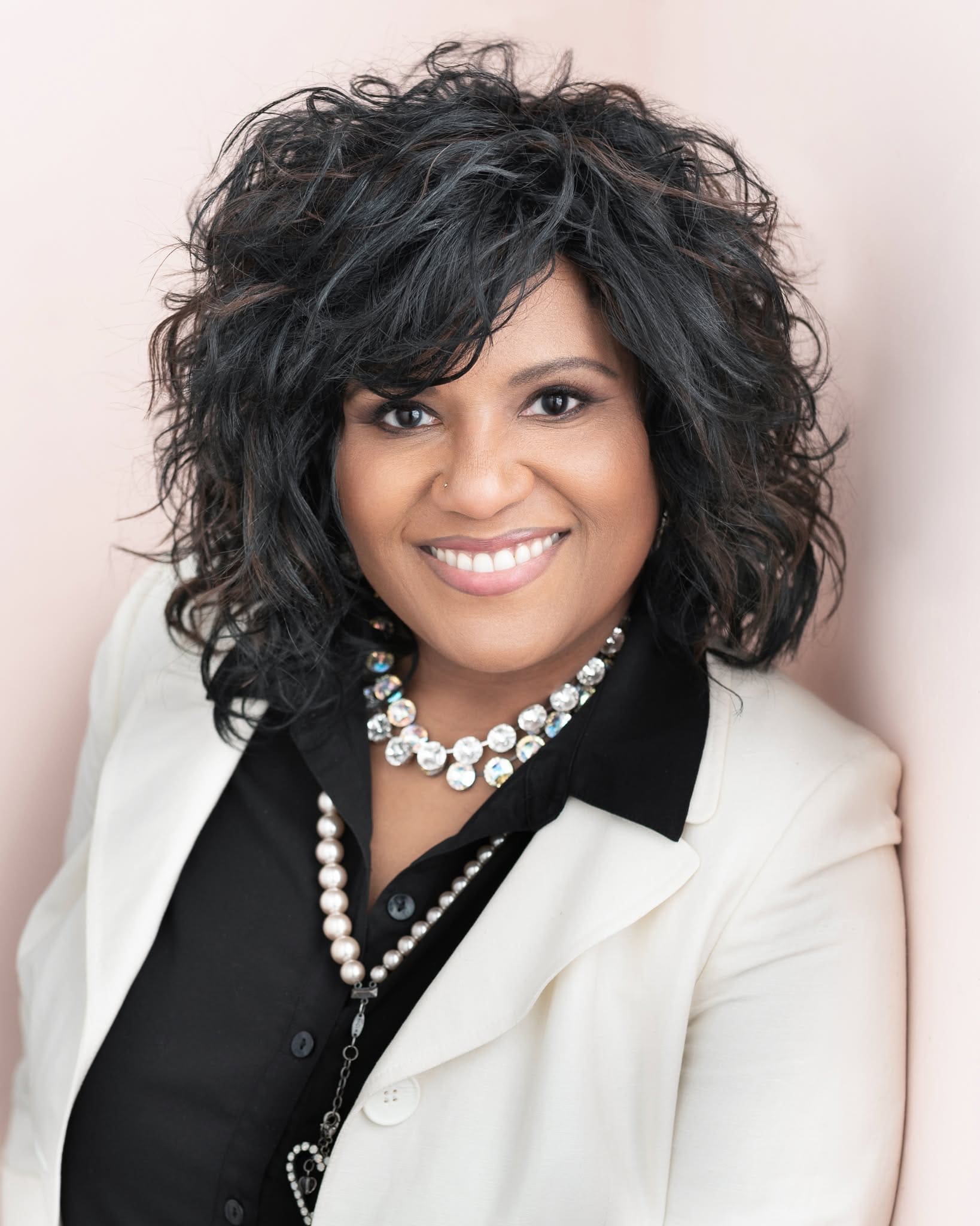 A smiling woman with voluminous dark curly hair wears a white blazer, black blouse, and layered crystal and pearl necklaces.