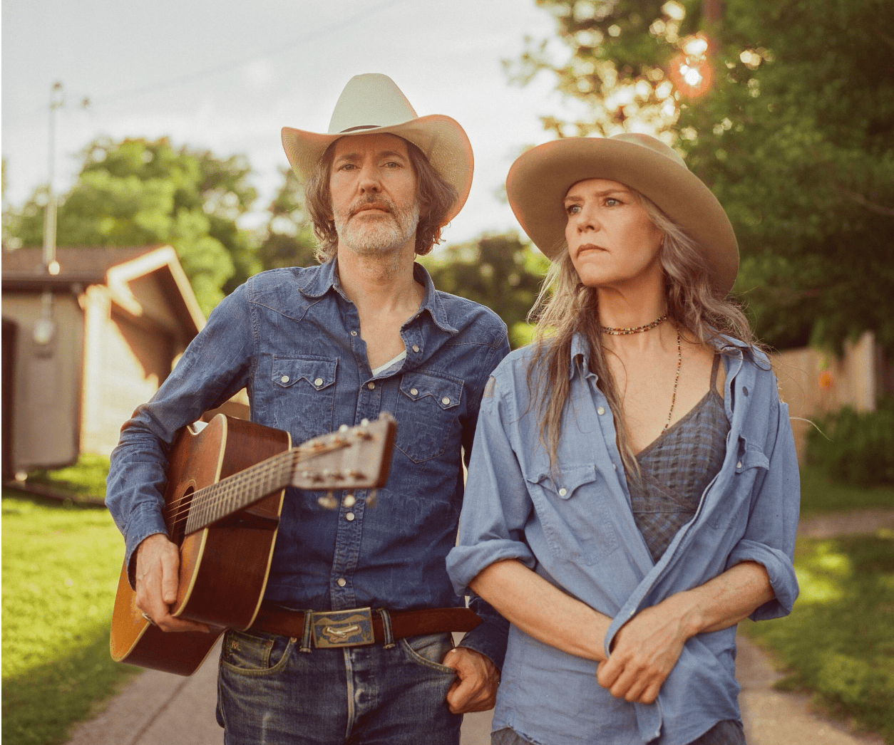 Two people in cowboy hats walk down a sunny tree-lined street; the man holds an acoustic guitar.