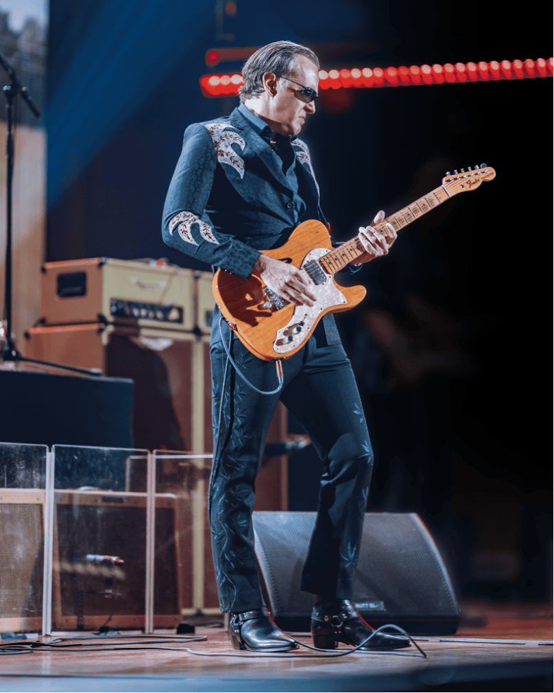 A guitarist in a dark embroidered western-style jacket and sunglasses plays a honey-colored Telecaster on a stage.