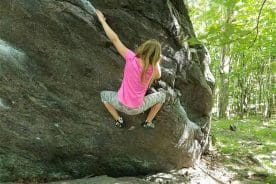 A climber in a pink shirt and gray patterned shorts scales a large boulder in a wooded area.